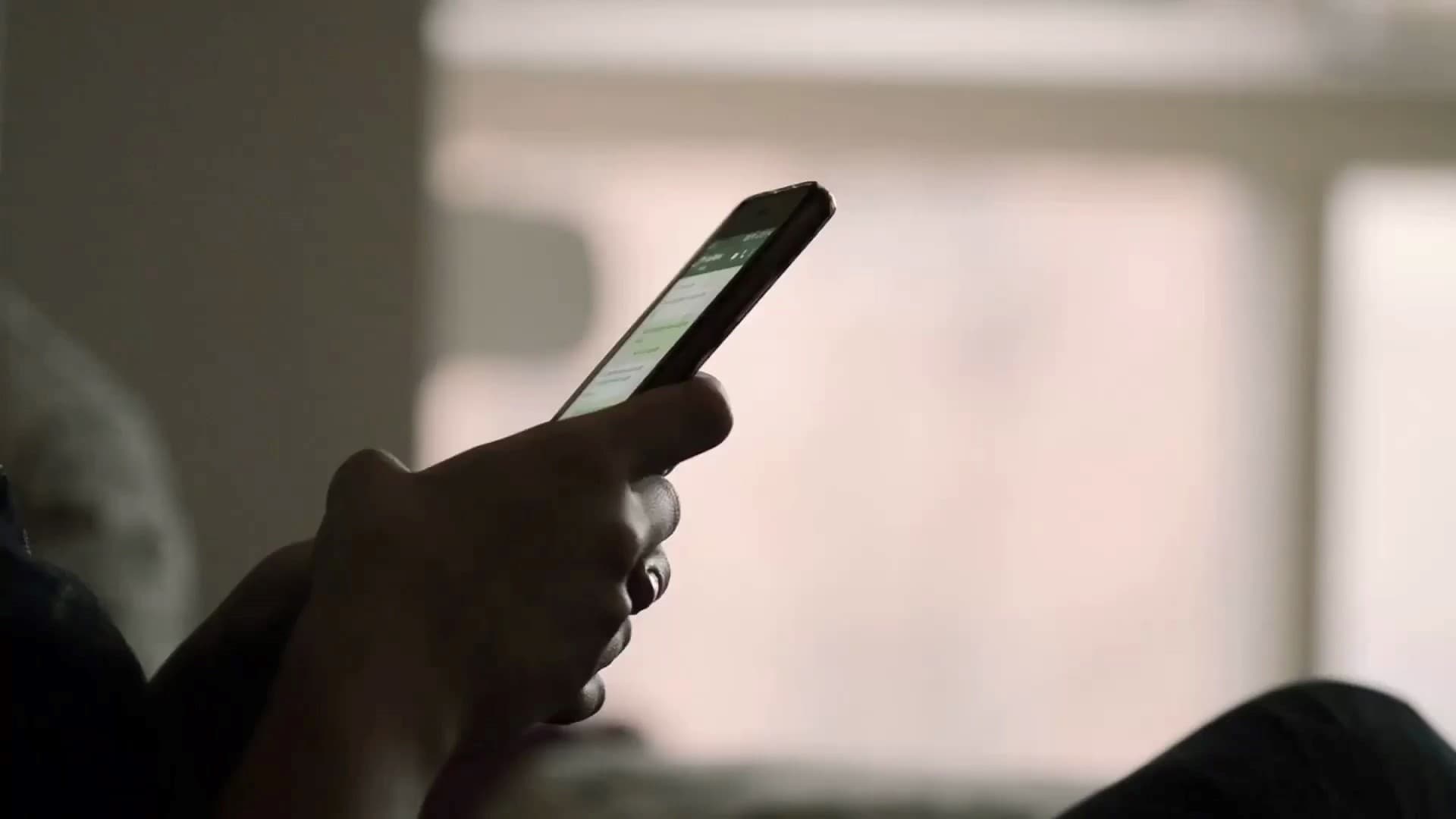 Person som holder en smarttelefon horisontalt, leser eller skriver på skjermen, med uskarp bakgrunn.