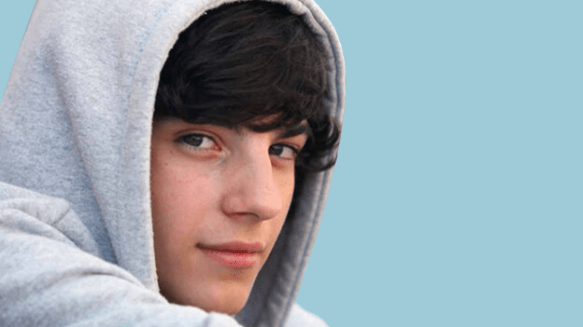 En ung person med mørkt hår og lys hud har på seg en grå hettegenser og ser seg litt over skulderen med et svakt smil mot en ensfarget lyseblå bakgrunn.