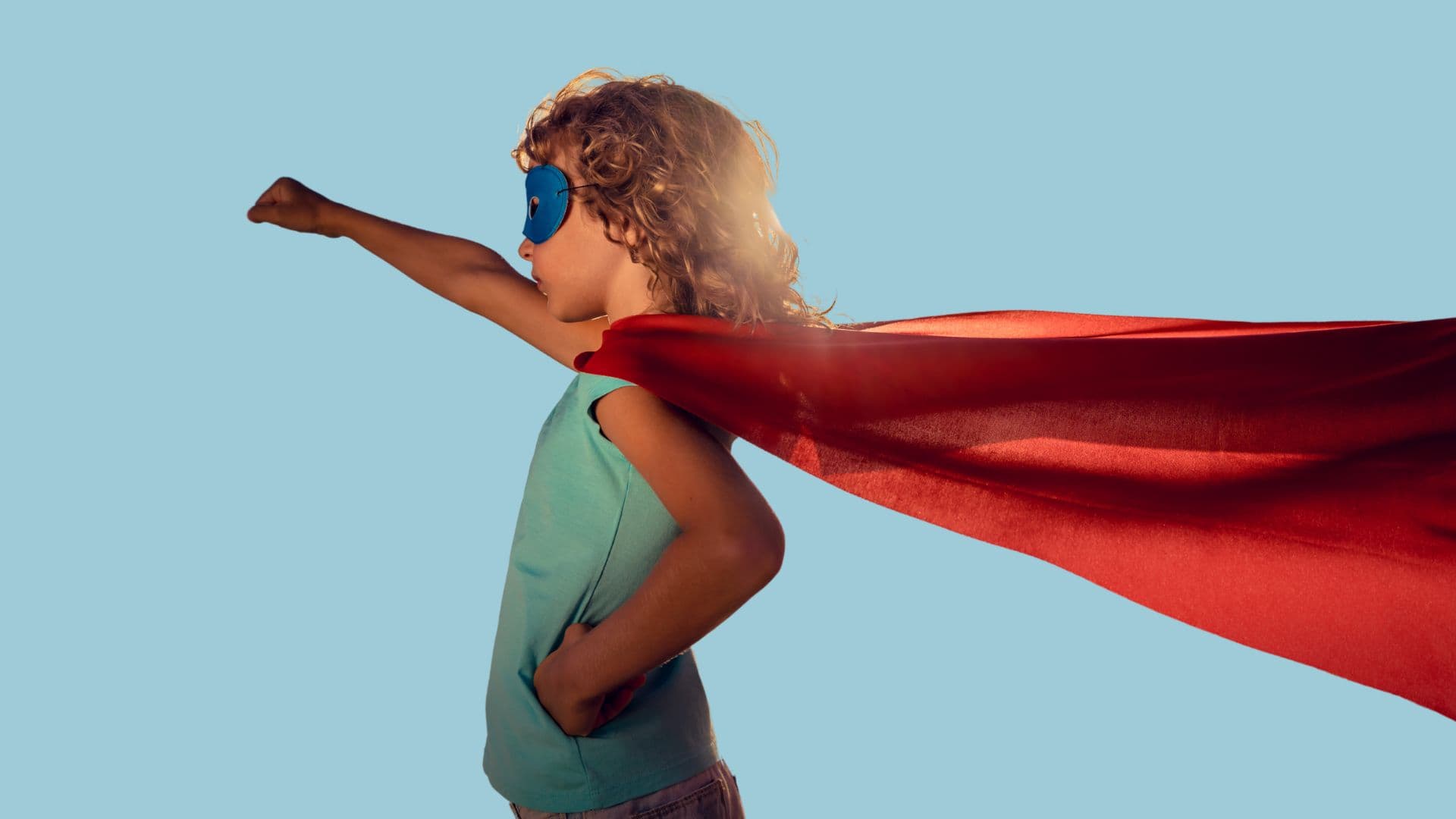 Et barn iført blå maske og rød kappe poserer som superhelt, stående i profil med den ene armen løftet og den andre på hoften, mot en ensfarget blå bakgrunn.