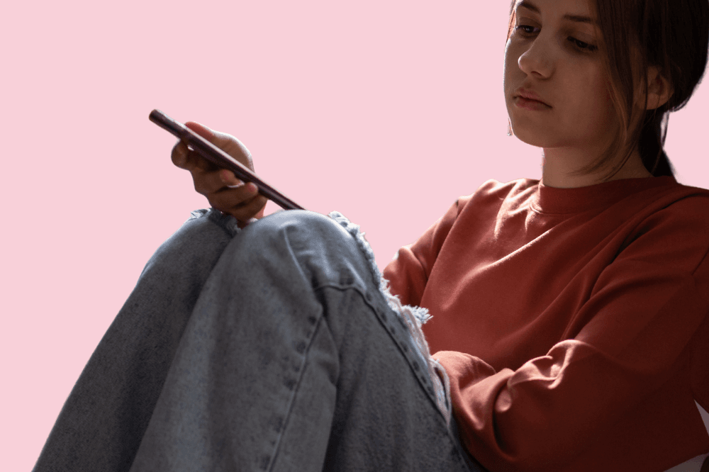 En ung kvinne i rustfarget genser og slitte jeans sitter med knærne i været og ser intenst på telefonen sin mot en ensfarget rosa bakgrunn.