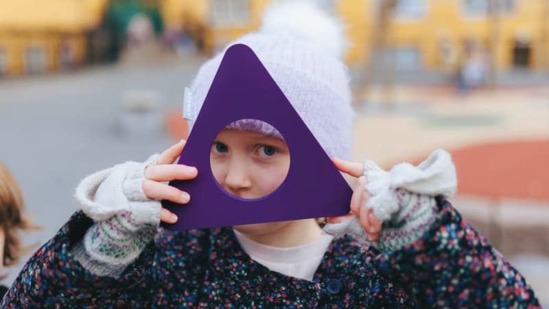 Et barn med en hvit strikkehatt holder en lilla, trekantet ramme med et rundt hull opp mot ansiktet sitt og ser gjennom den. Bakgrunnen er uskarp og viser et uteområde med gule bygninger.