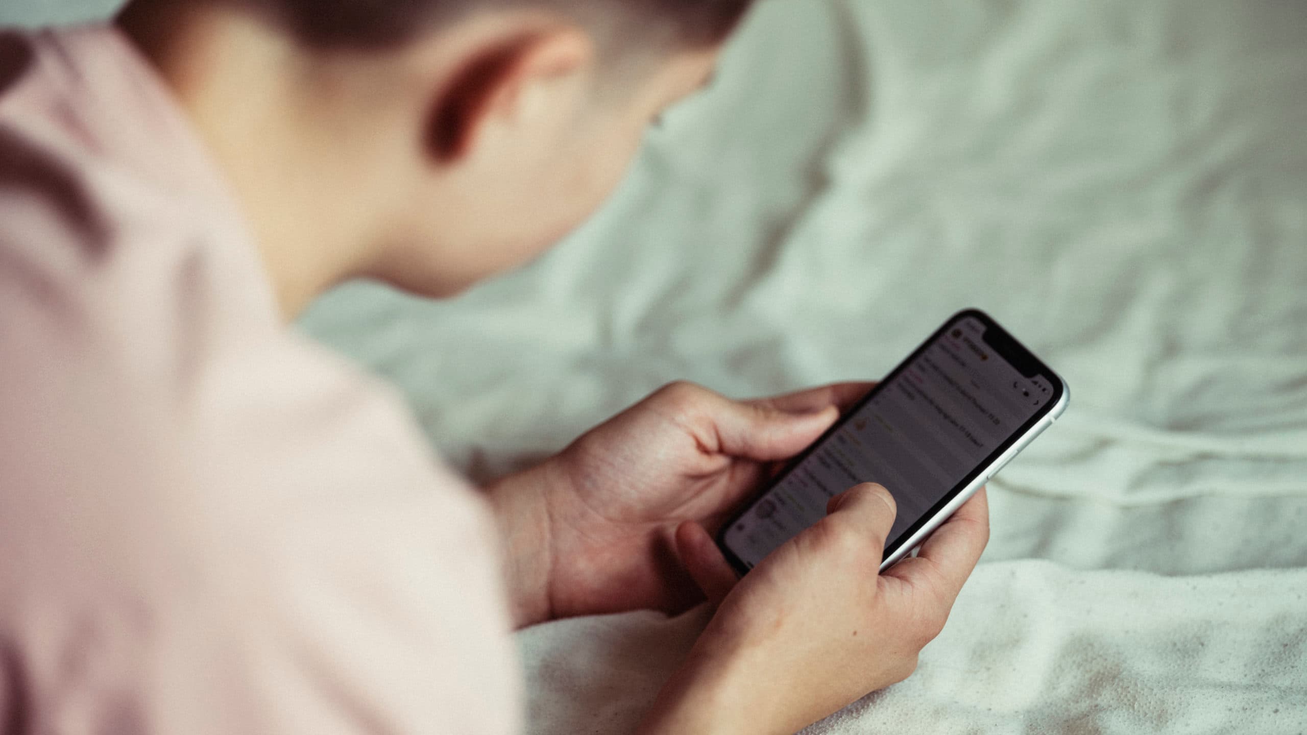 En person ligger på en seng og bruker en smarttelefon mens han leser eller skriver meldinger i en meldingsapp. Bildet er fokusert på telefonen og hendene, mens ansiktet ikke er synlig.