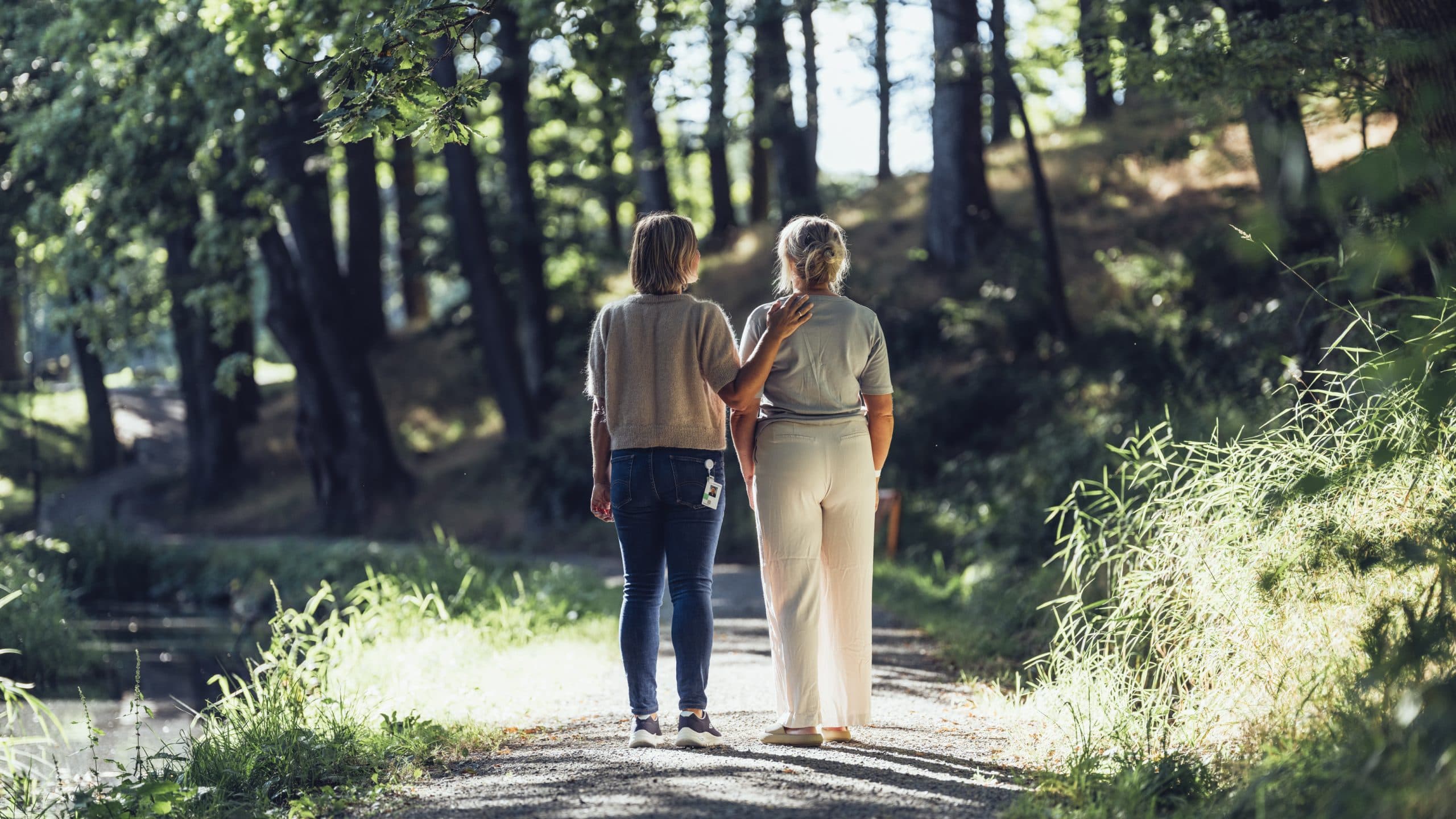 To personer som går på en skogssti, den ene trøster den andre med en hånd på skulderen, omgitt av trær og gress.