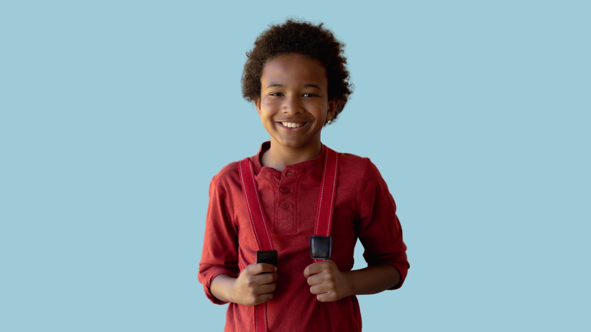 Smilende barn med krøllete hår, iført rød langermet skjorte og med røde bukseseler, mot en ensfarget lyseblå bakgrunn.