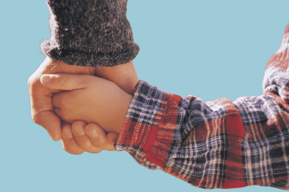 Nærbilde av to personer som holder hverandre i hånden mot en lyseblå bakgrunn. Den ene personen har på seg et grått ullærme, mens den andre har på seg en rød og blå rutete skjorte.