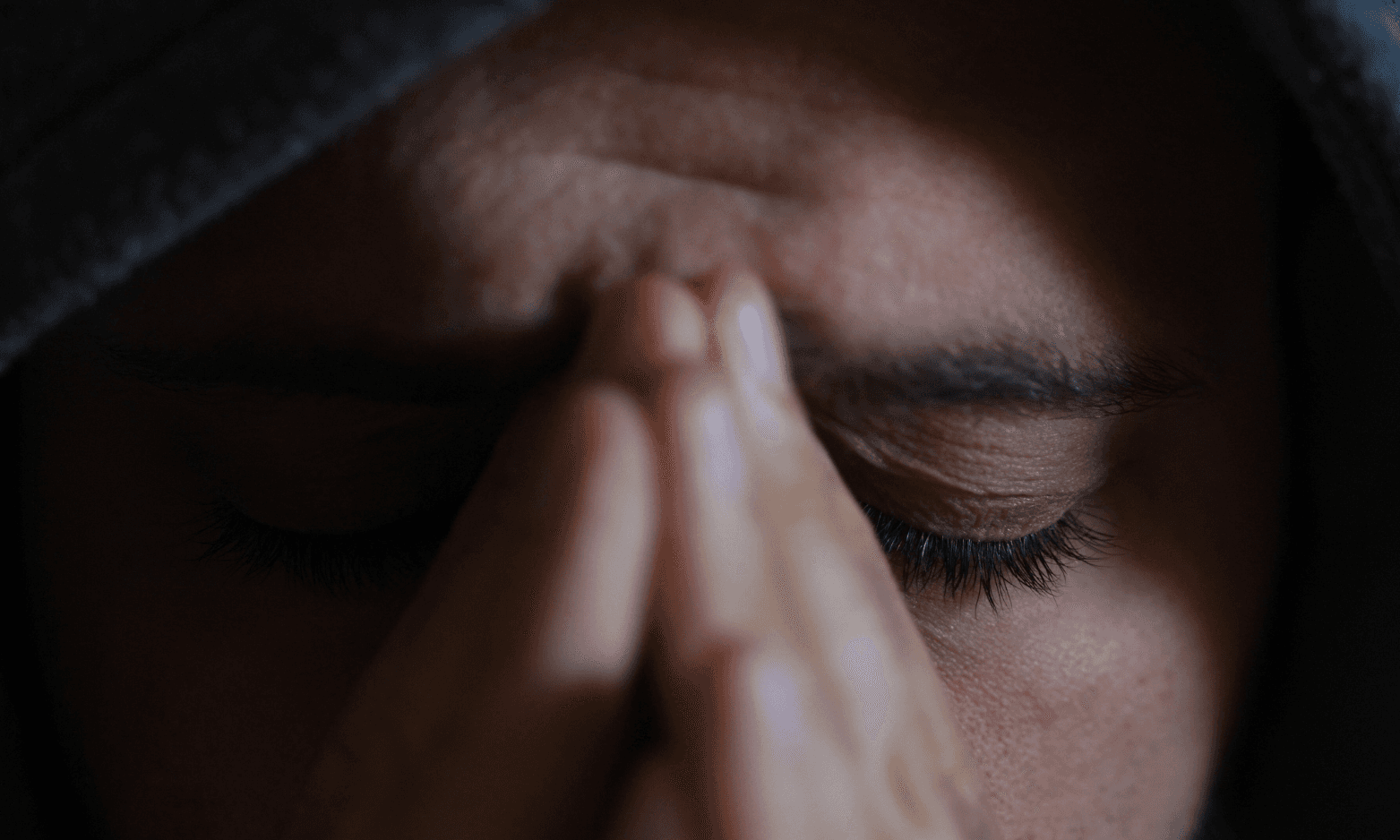 Nærbilde av en person med lukkede øyne som presser hendene sammen mot ansiktet som om hun ber eller er i dype tanker, delvis tildekket av en hette.