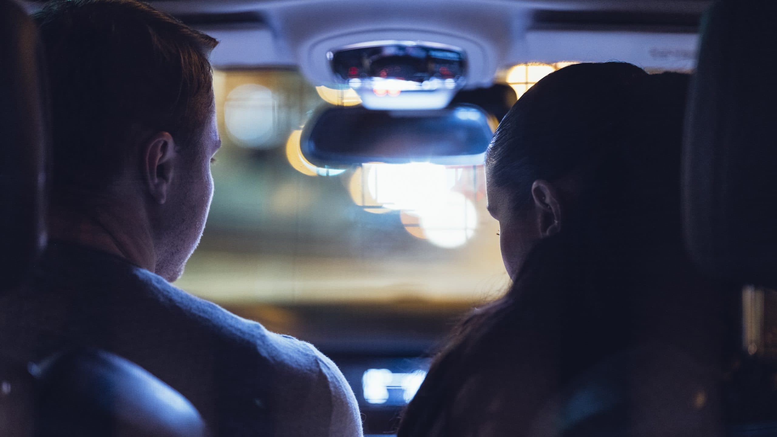 En mann og en kvinne sitter i forsetene i en bil om natten, sett bakfra. Byens lys er uskarpt i bakgrunnen gjennom frontruten.